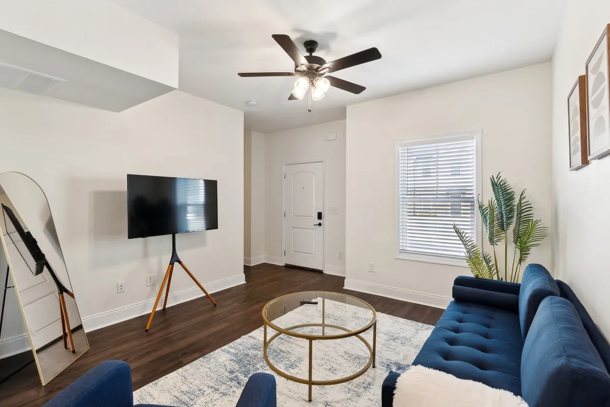 This image showcases a navy blue sofa, glass coffee table, large mirror, television on a stand, and a lush indoor plant. The setting is arranged for comfort and socialization, inviting guests to relax, watch TV, or settle in for a conversation. The room is tastefully decorated and exudes a cozy, inviting vibe.