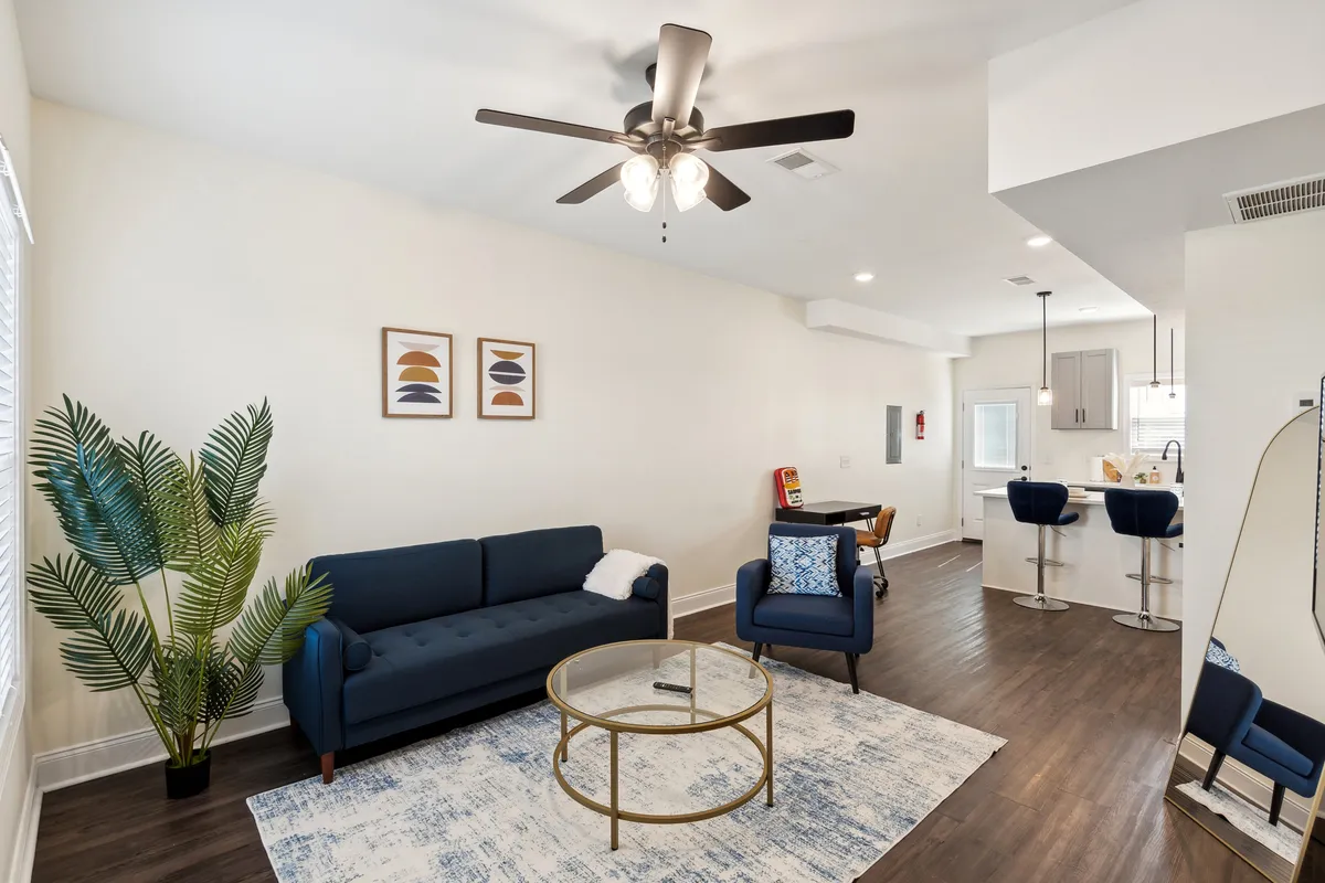 A comfortable living space featuring a sofa, coffee table, stylish artwork, a plant, and adjacent bar stools. The area is open and bright, blending spaces for both relaxation and entertaining. The modern decor and layout encourage sociability and connection.