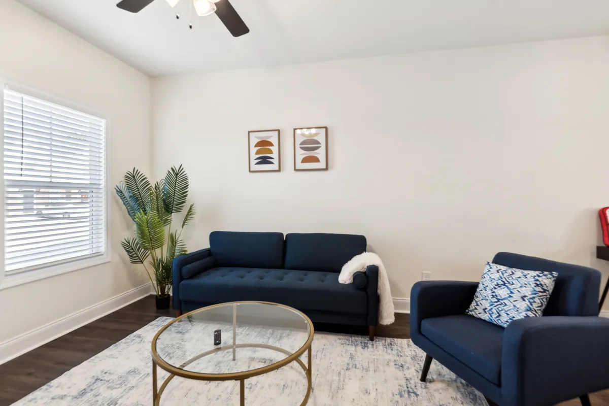 The image showcases a modern, inviting living space featuring a plush blue sofa, a sleek glass coffee table, vibrant wall art, and a lush indoor plant. Morning light gently fills the room, creating a warm and welcoming atmosphere perfect for relaxation or gathering with family.