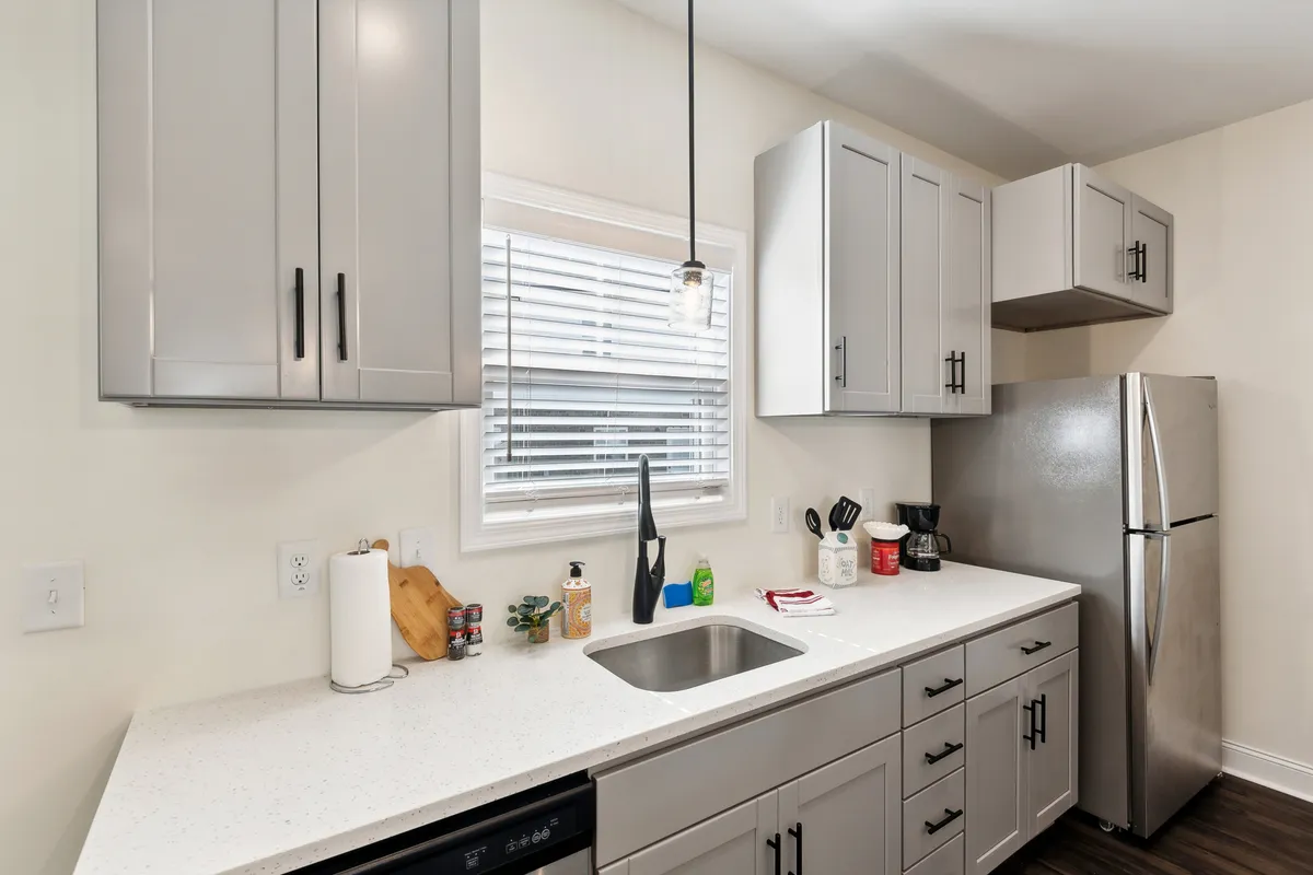 A bright and functional kitchen countertop scene, with a sink, window, and a view of the refrigerator. The space feels open and practical, ideal for everyday cooking and social moments like making morning coffee or preparing meals together.