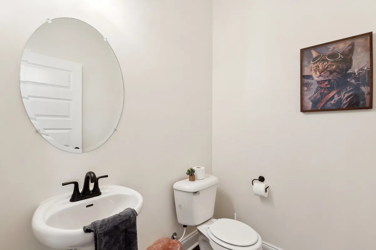 A well-lit powder room featuring a round mirror above a modern sink, accented by a whimsical cat portrait on the wall and a soft lavender towel hanging nearby. The space exudes a cheerful and inviting vibe, perfect for guests' comfort.