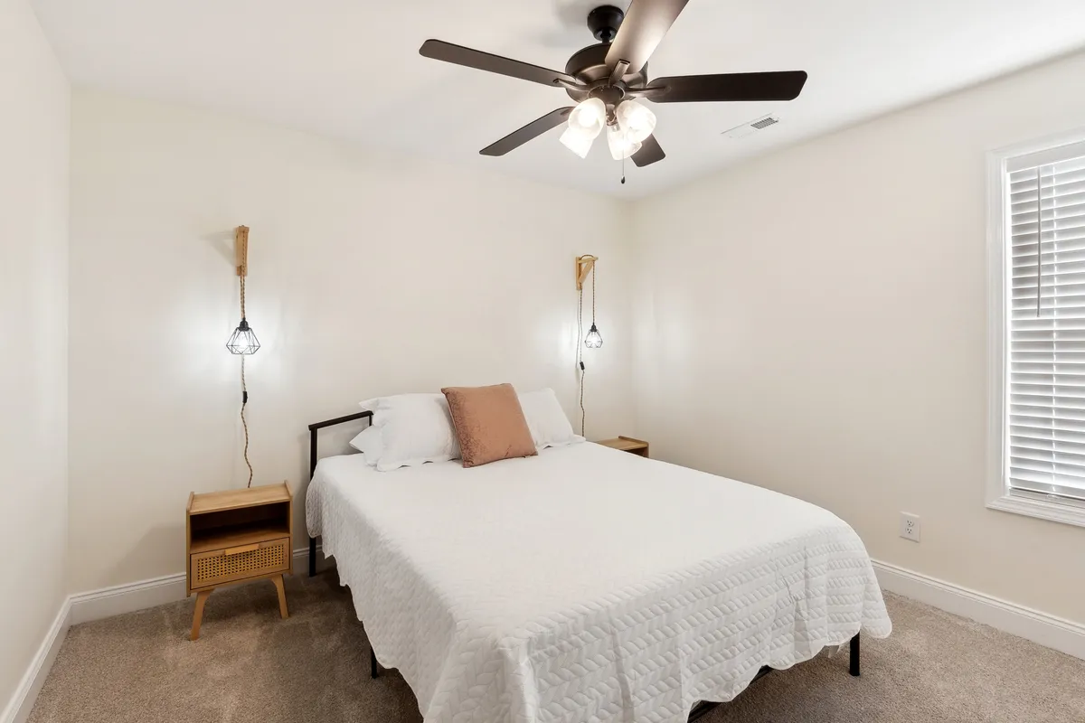 A tranquil bedroom scene featuring a bed with a white quilt, nightstands with softly glowing lamps, and a ceiling fan above. Natural light gently filters into the room, creating a serene atmosphere ideal for rest and relaxation.